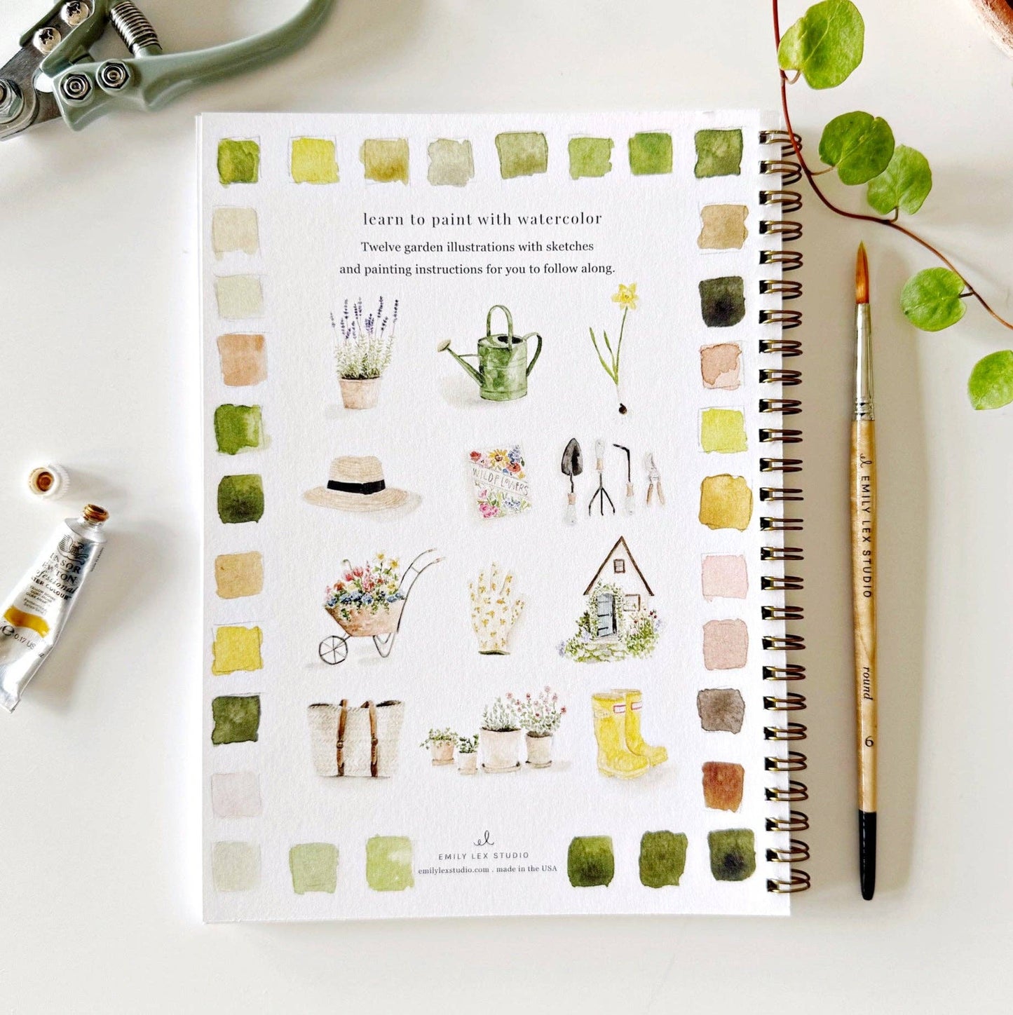 Notebook with watercolor illustrations of garden-themed designs on a white surface with art supplies.