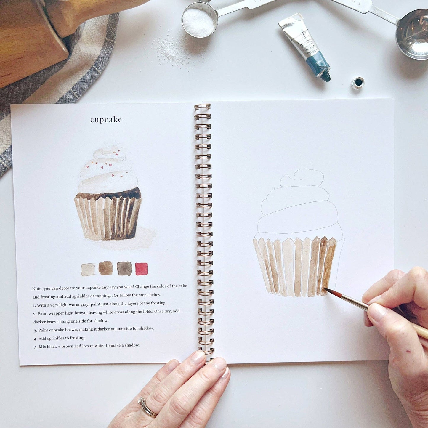 Open book with cupcake illustration and hands using a paintbrush