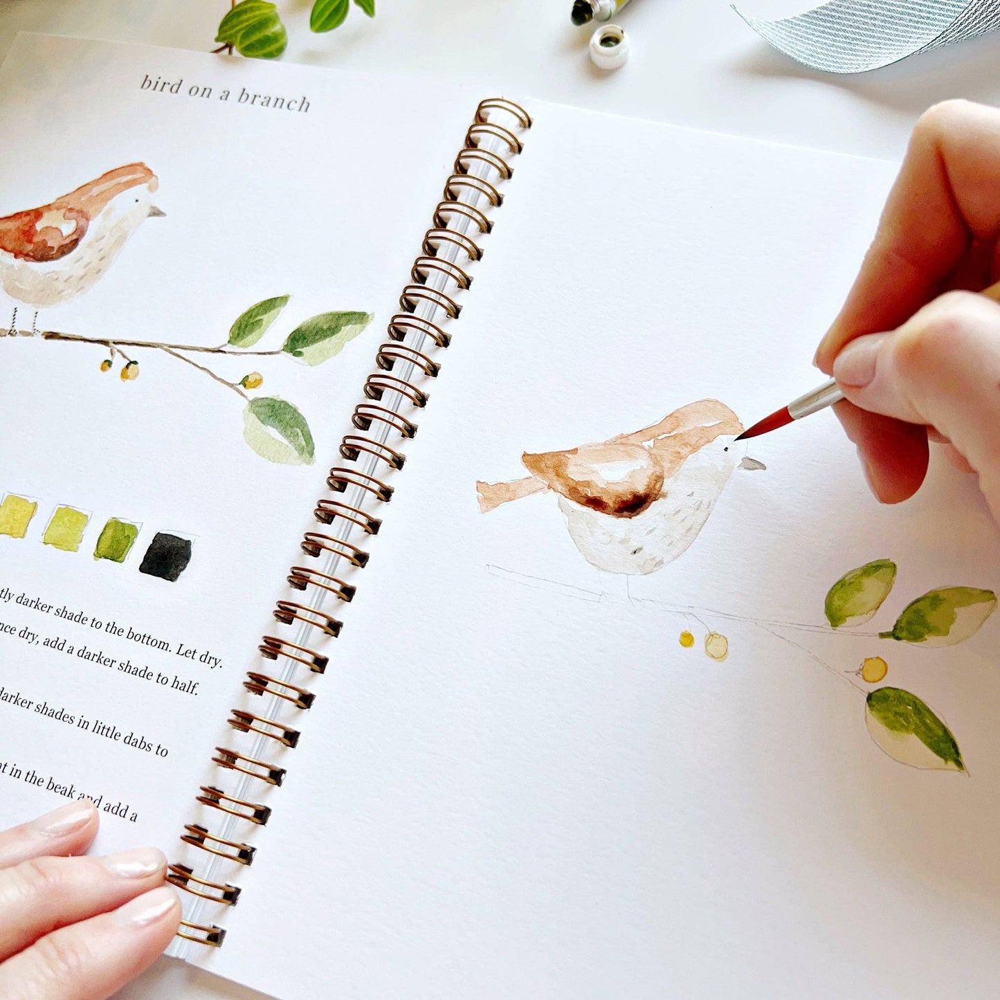 Person painting a bird illustration in a notebook with a paintbrush.
