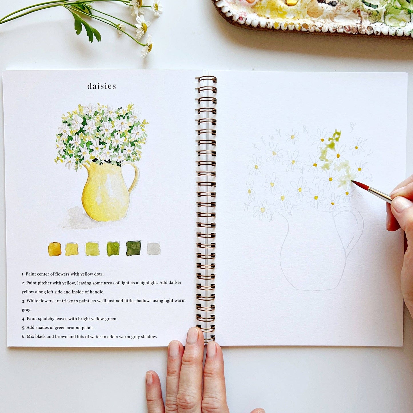 Open art book with a painting of daisies and a hand holding a paintbrush.