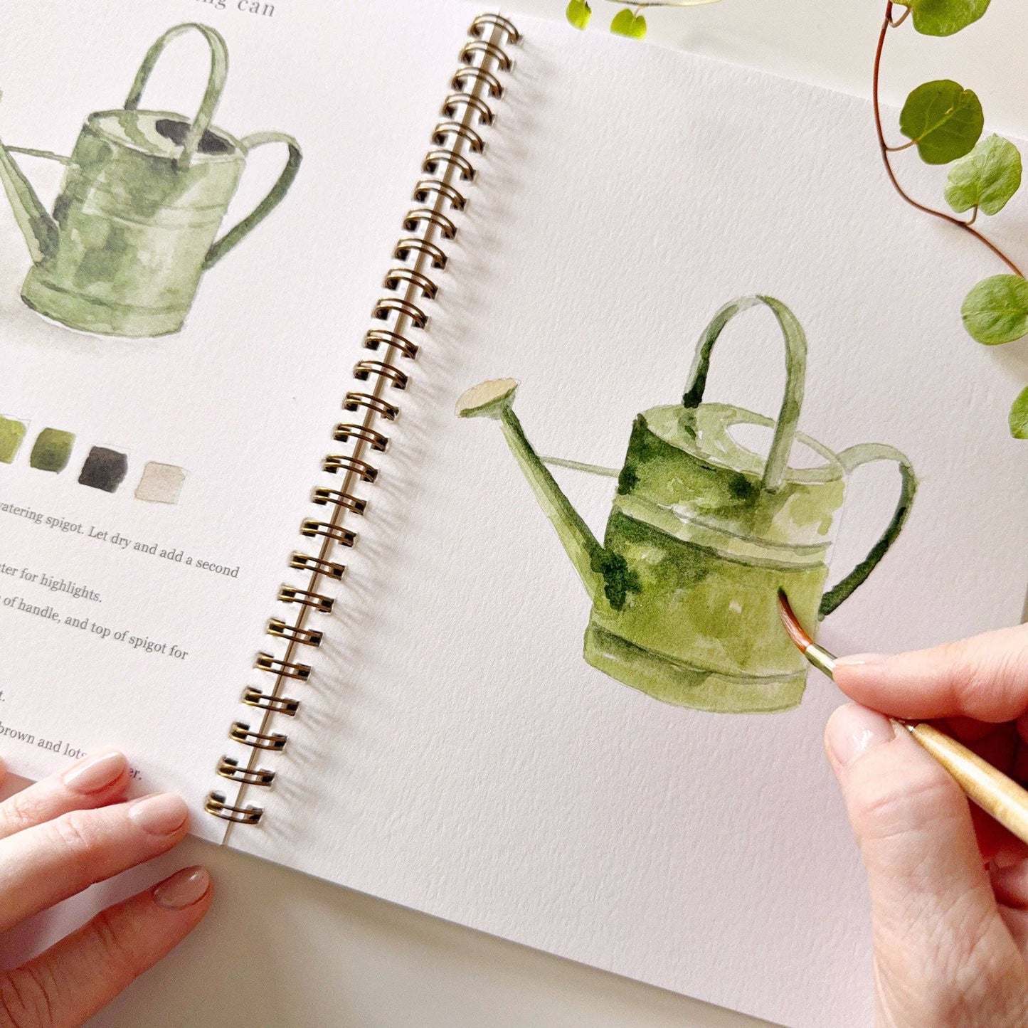 Person drawing a green watering can in a notebook with a real watering can and leaves on the side.