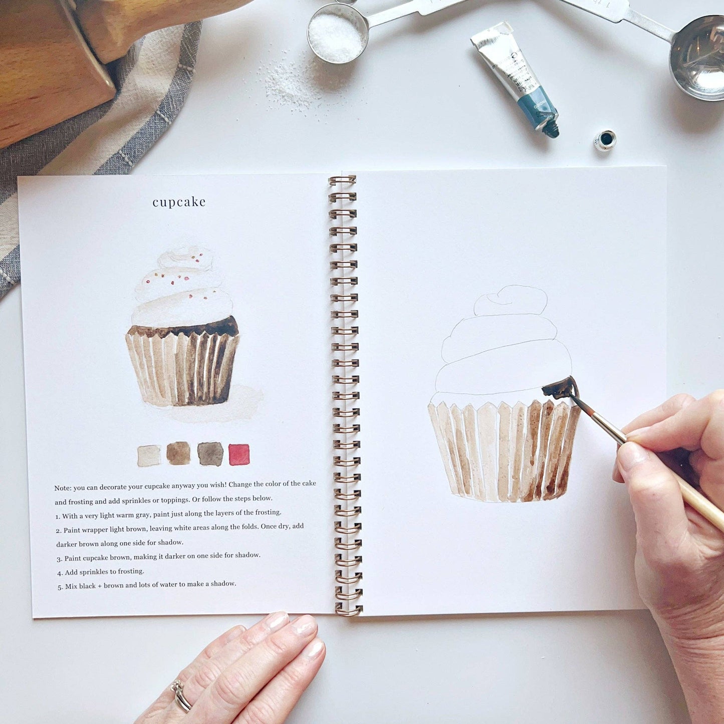 Open book with cupcake illustration and text, hand holding a brush near the illustration.