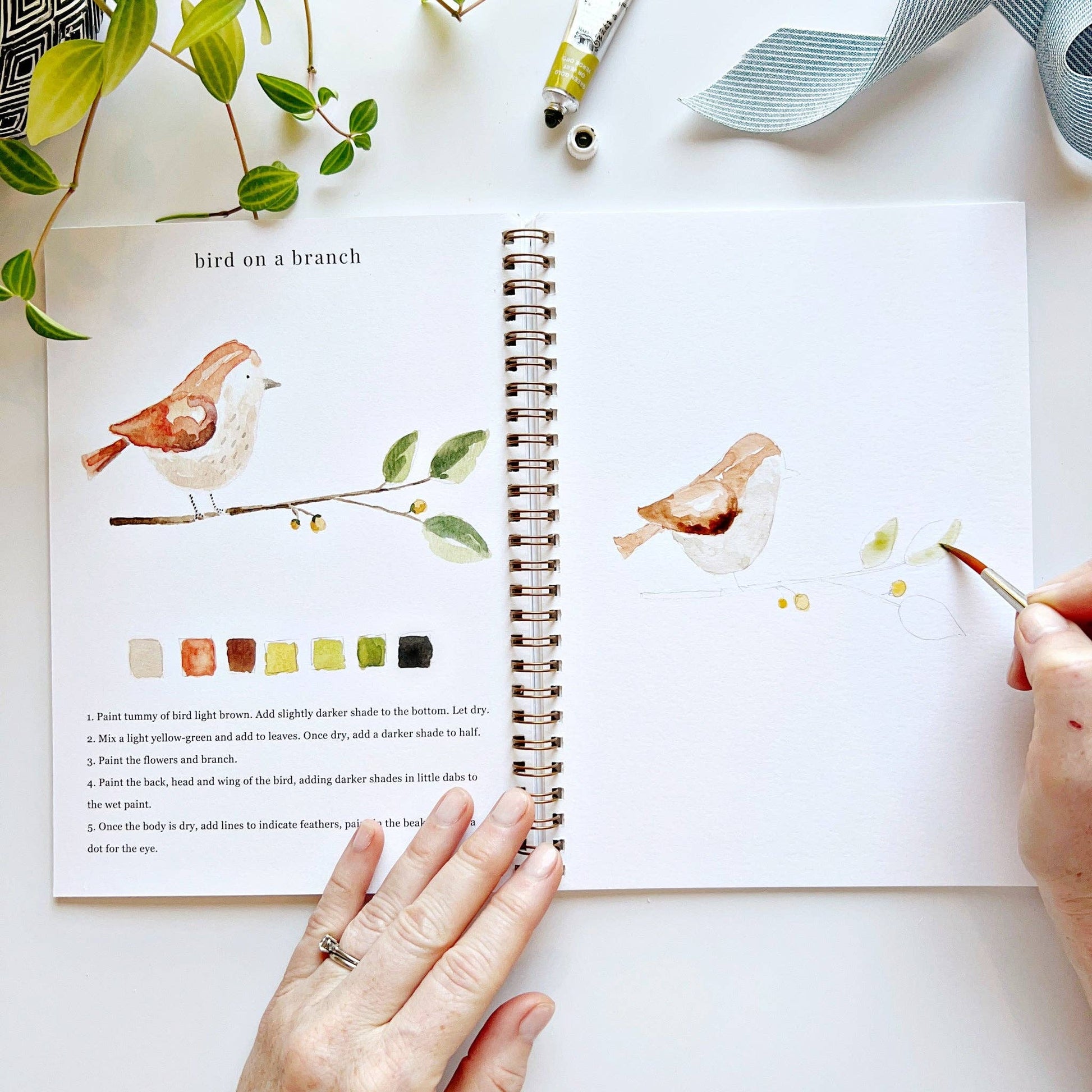 Artistic depiction of a bird on a branch with watercolor painting supplies and a hand holding a brush.
