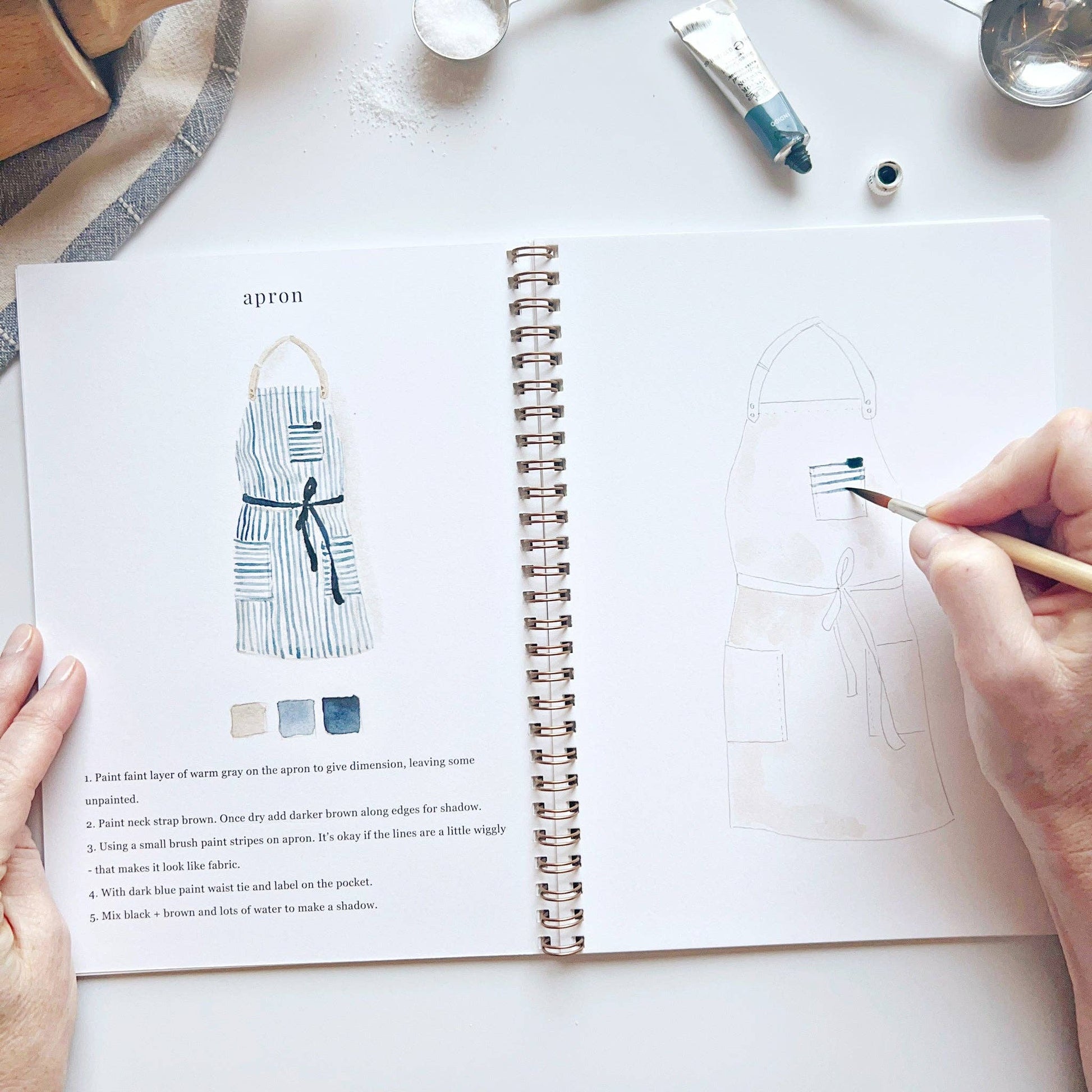 Person holding a paintbrush over an open book with apron design illustrations and instructions.