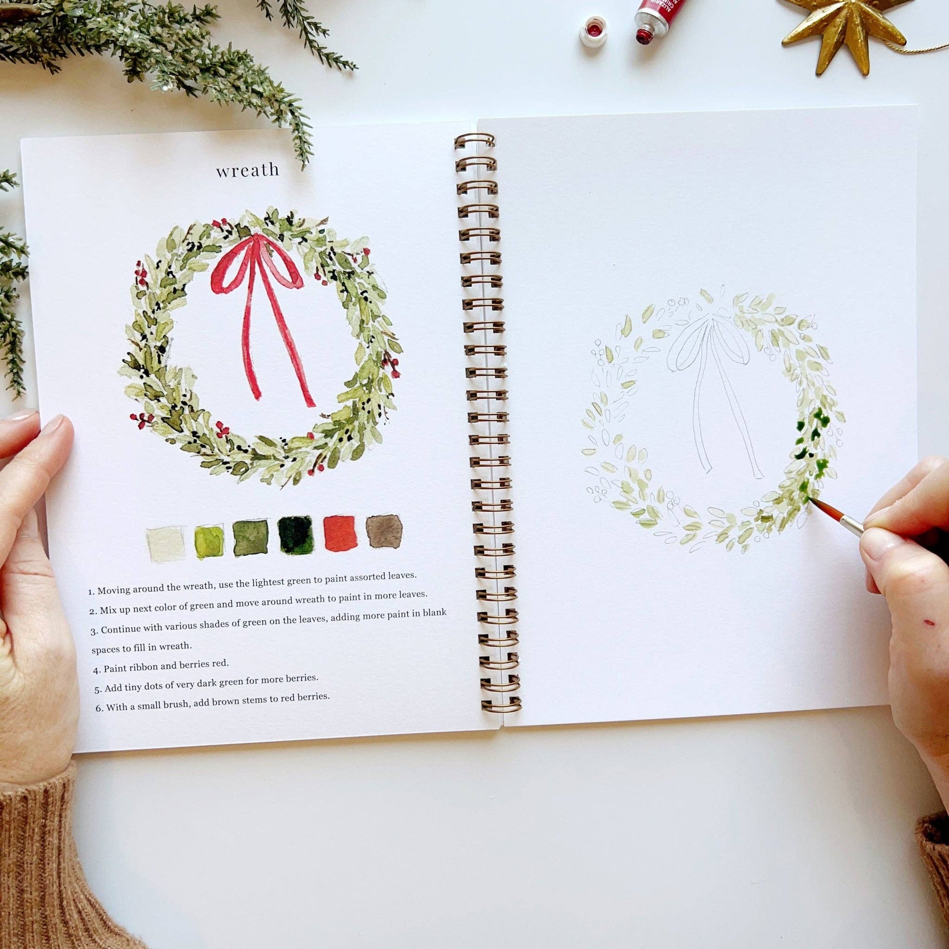 Open book with drawing of a wreath and color swatches, surrounded by Christmas decorations.