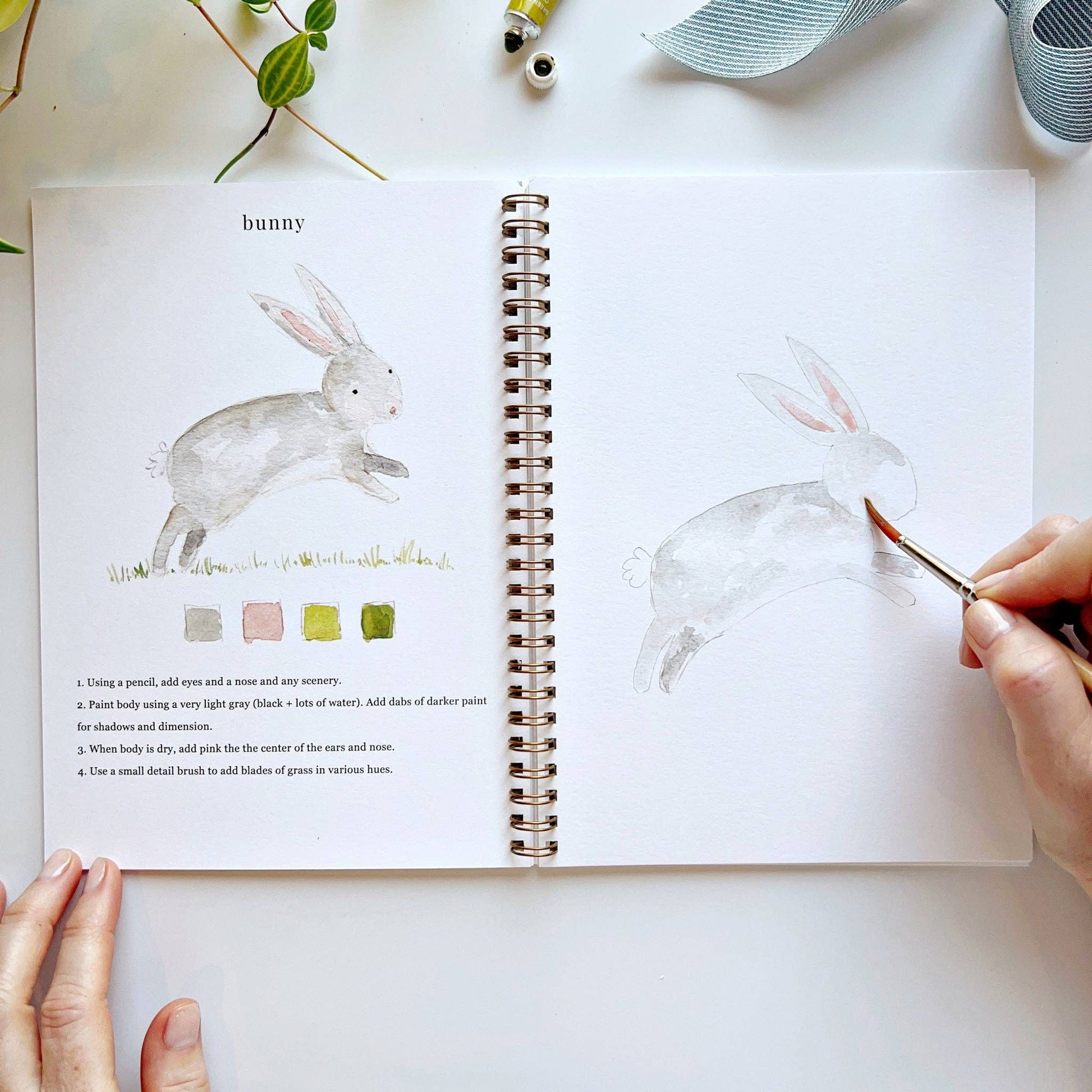 Open book with watercolor illustrations of a bunny, being painted by a hand with a brush.